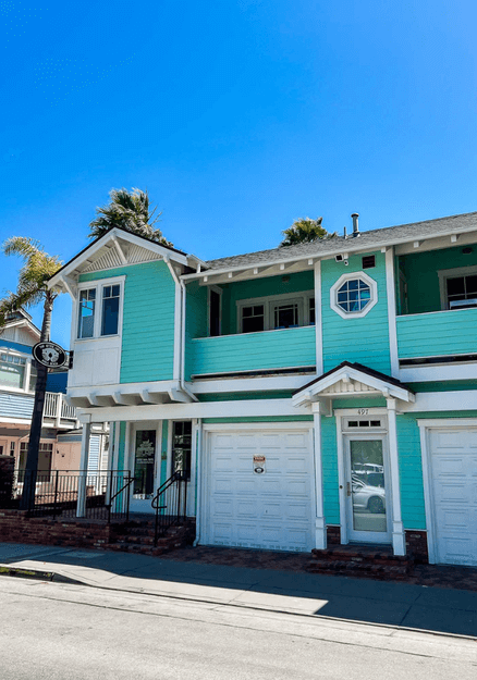 A charming teal green beach cottage at Avila Beach, featuring a cozy porch with white trim, surrounded by vibrant plants and overlooking the peaceful seaside, with the ocean and clear sky visible in the background.